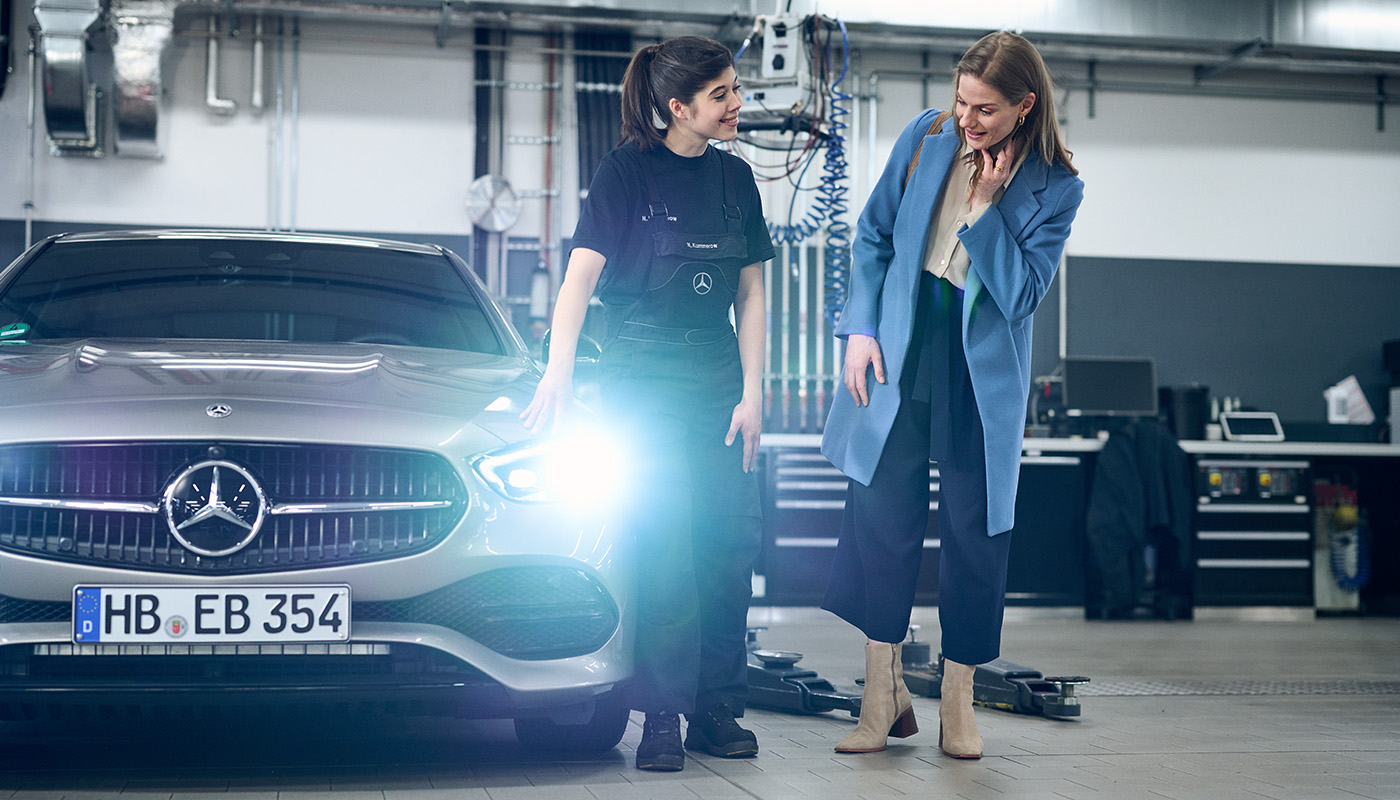 Eine Kundin steht mit einer Werkstattmitarbeiterin in der Werkstatt und inspizieren gemeinsam die angeschaltetet Frontbeleuchtung eines silbernen Mercedes