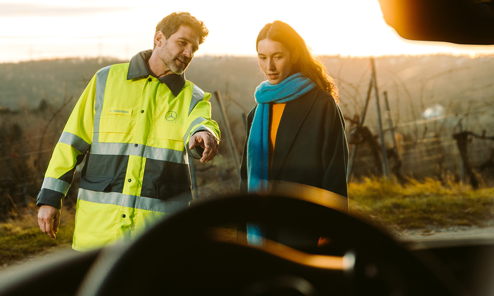 Mann vom Mercedes-Benz Pannenservice steht mit der Fahrerin vor ihrem Mercedes und weist auf den Schaden hin